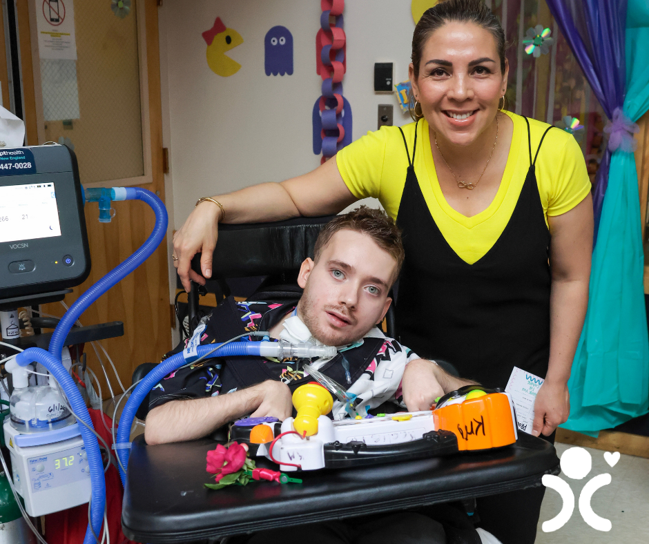 A woman smiles beside a young man in a wheelchair with medical equipment in a colorful room.