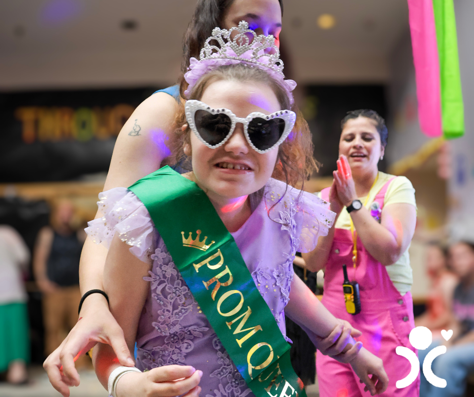 Smiling girl in a prom queen sash, tiara, and heart glasses is supported by a woman at a joyful event.