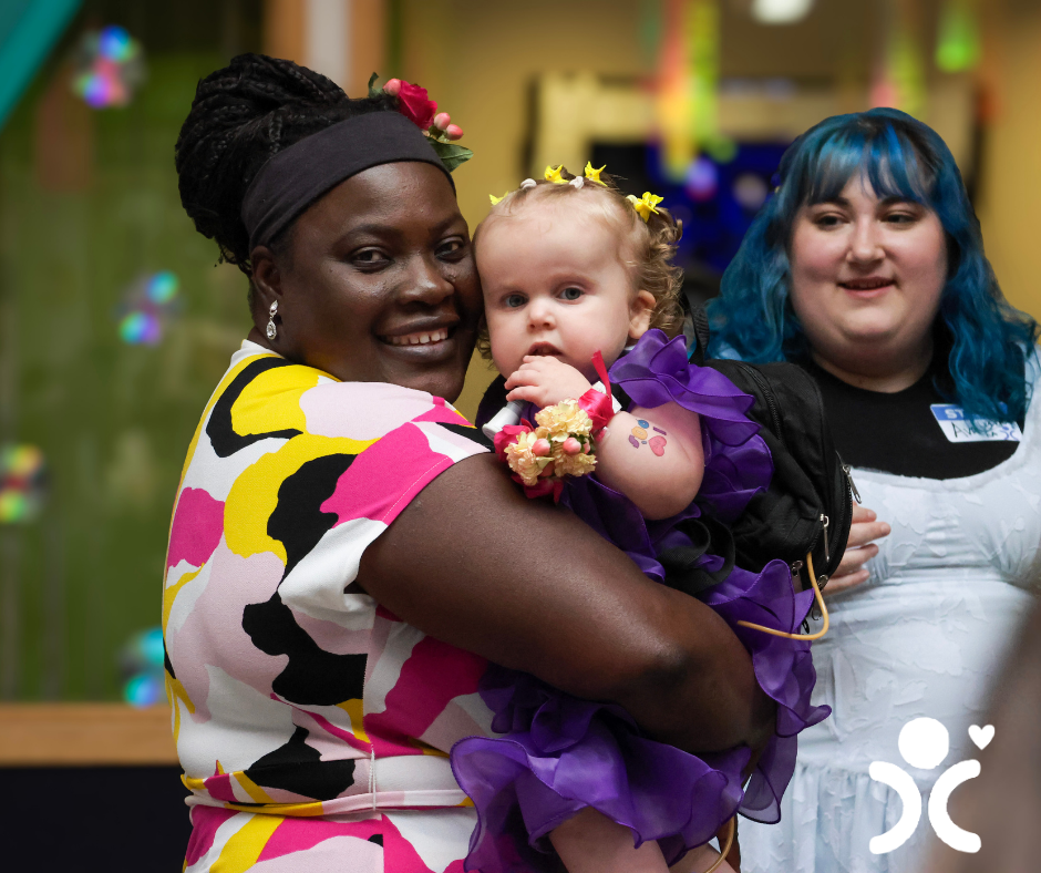 Woman in a colorful dress holds a baby in purple; another woman with blue hair stands behind them, smiling.