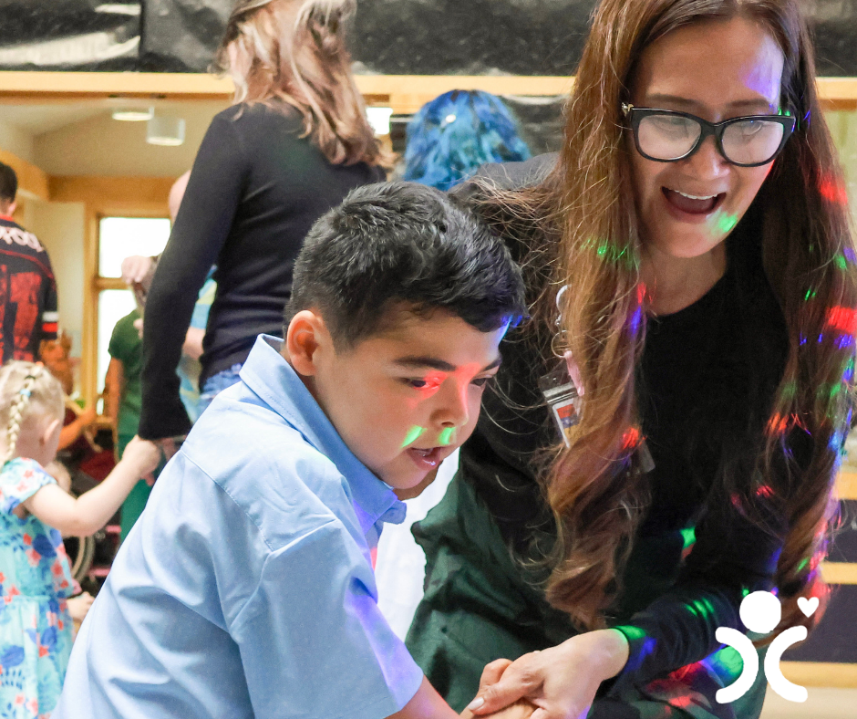 A smiling woman helps a young boy at a lively indoor event with colorful light spots on their faces.