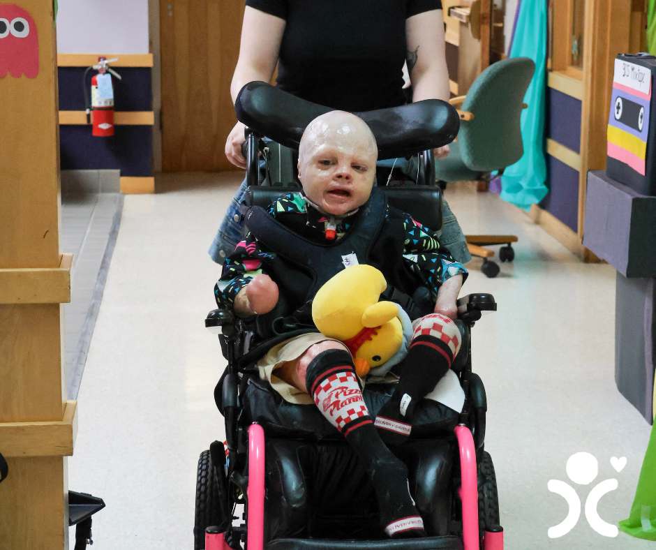 A child with bandages sits in a wheelchair holding a stuffed toy, being pushed by an adult in a hallway.