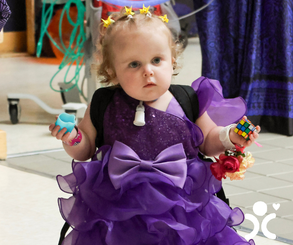 Toddler in a ruffled purple dress holds toys, with medical gear on neck and star clips in curly hair.