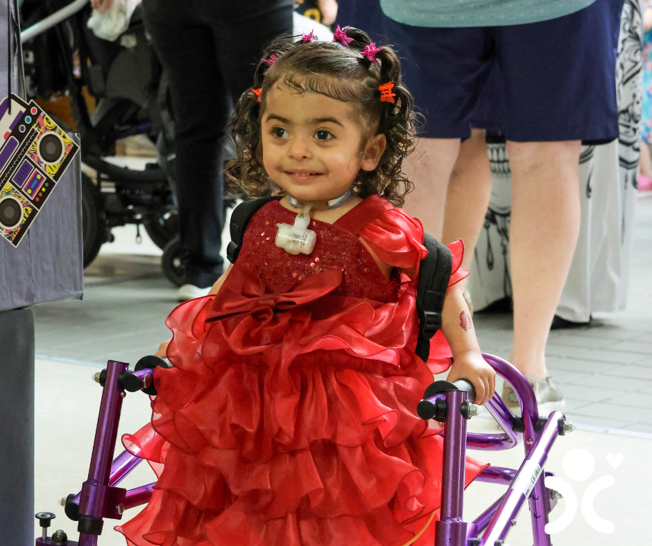 Smiling young girl in a red dress using a purple walker indoors, with people walking in the background.