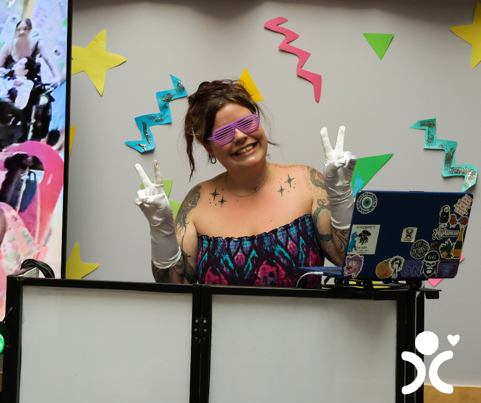 Smiling woman in sunglasses and gloves makes peace signs at a decorated DJ booth with colorful shapes.