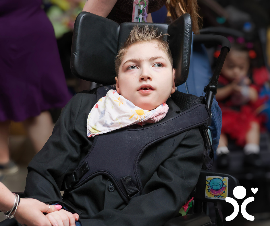 A boy in a wheelchair wears a suit and bib, holding hands with someone. A logo is in the corner.