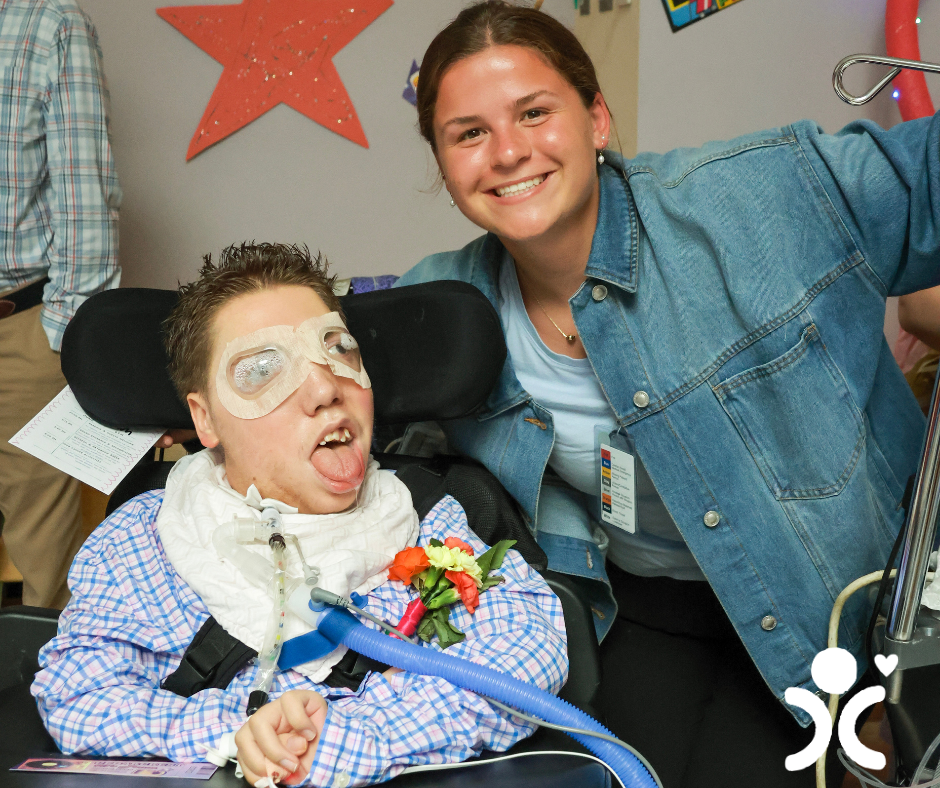 Smiling woman stands next to a man in a wheelchair wearing a boutonniere and medical equipment.