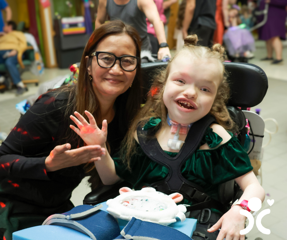 Woman smiling with a young girl in a wheelchair, both happy at an indoor event. Accessible inclusion symbol shown.