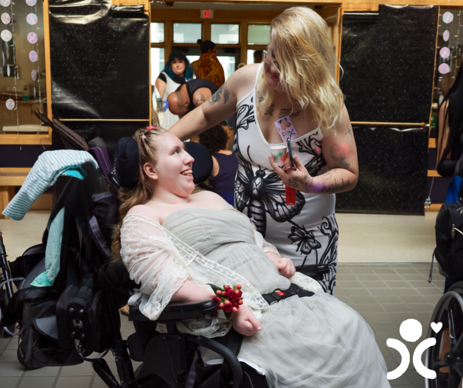 Woman in a wheelchair smiles at another woman fixing her hair in a decorated indoor setting.