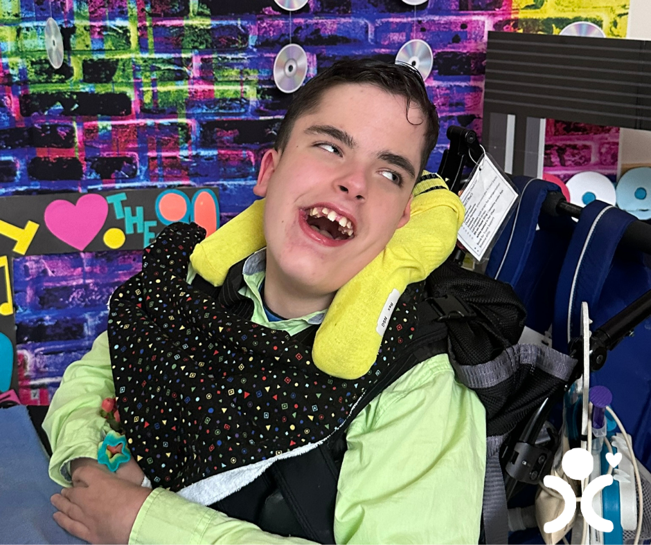 Smiling boy in a wheelchair with a colorful background and a yellow pillow around his neck.