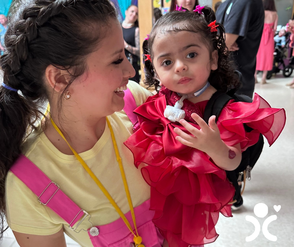 Smiling woman holds a young girl in a bright red ruffled dress at a busy indoor event.