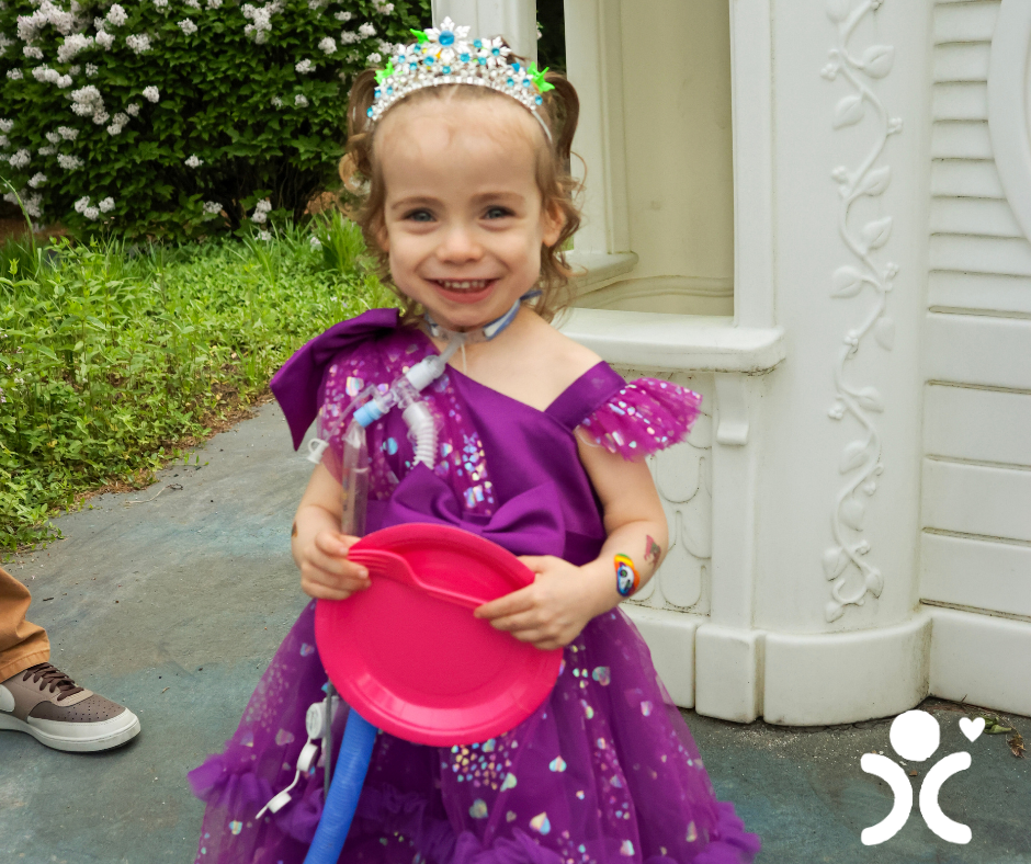 Smiling child in a purple princess dress holds a pink plate, standing outside near a white playhouse.