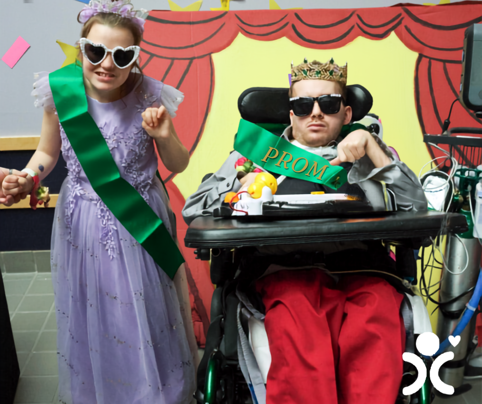 Two people in prom attire and green sashes pose, one in a wheelchair. Cartoon backdrop and prom crown visible.