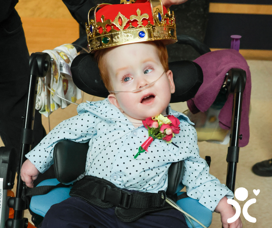 A child in a wheelchair wears a crown and boutonniere, smiling with a breathing tube.