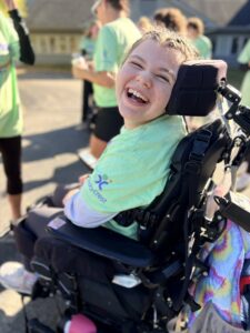 A smiling person in a light green shirt sits in a wheelchair outdoors.