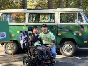 A young man in a wheelchair and a smiling man kneeling beside him in front of a green vintage VW bus.