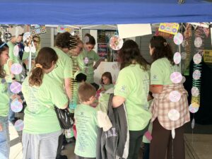 A group of people in matching light green shirts stand under a blue canopy decorated with hanging CDs and 90s imagery.