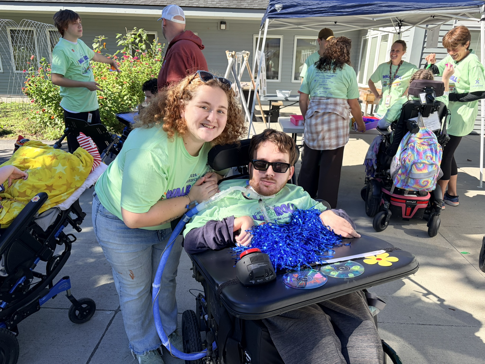 A smiling young woman stands behind a young man in a wheelchair with a blue pom-pom on his lap tray.