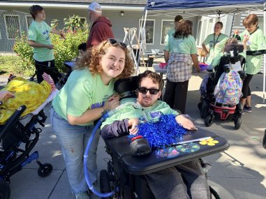 A smiling young woman stands behind a young man in a wheelchair with a blue pom-pom on his lap tray.